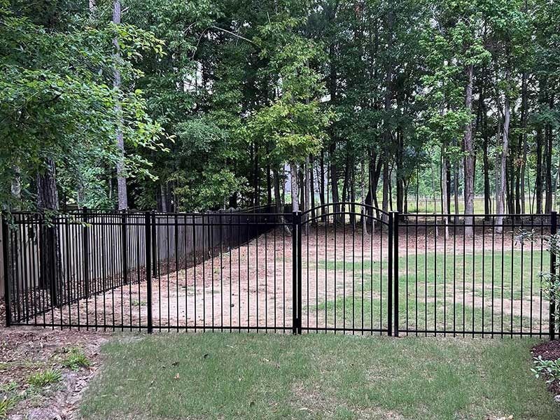 Black metal fence in backyard, separating grass from woods, under a cloudy sky.