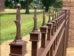 Brown metal fence with decorative finials, against a grassy area and brick wall.