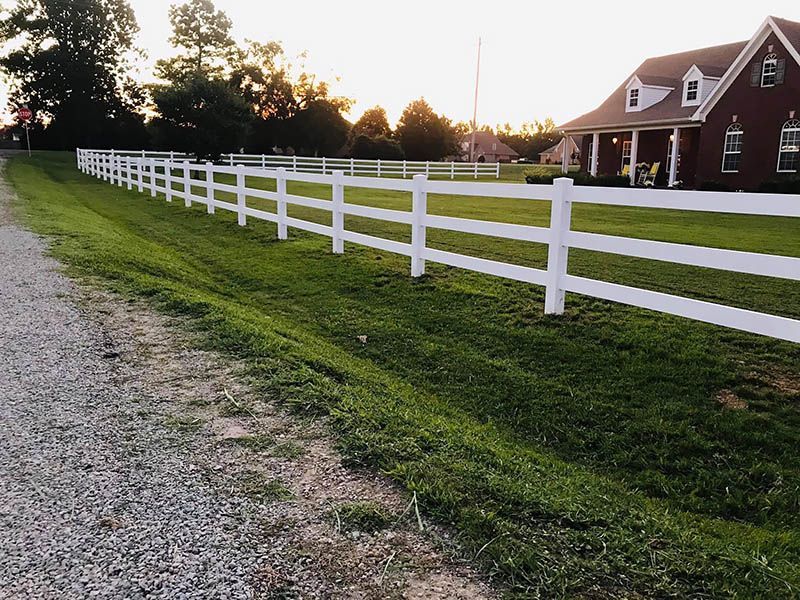 White fence bordering a grassy yard and gravel drive, in front of a brick house at dusk.