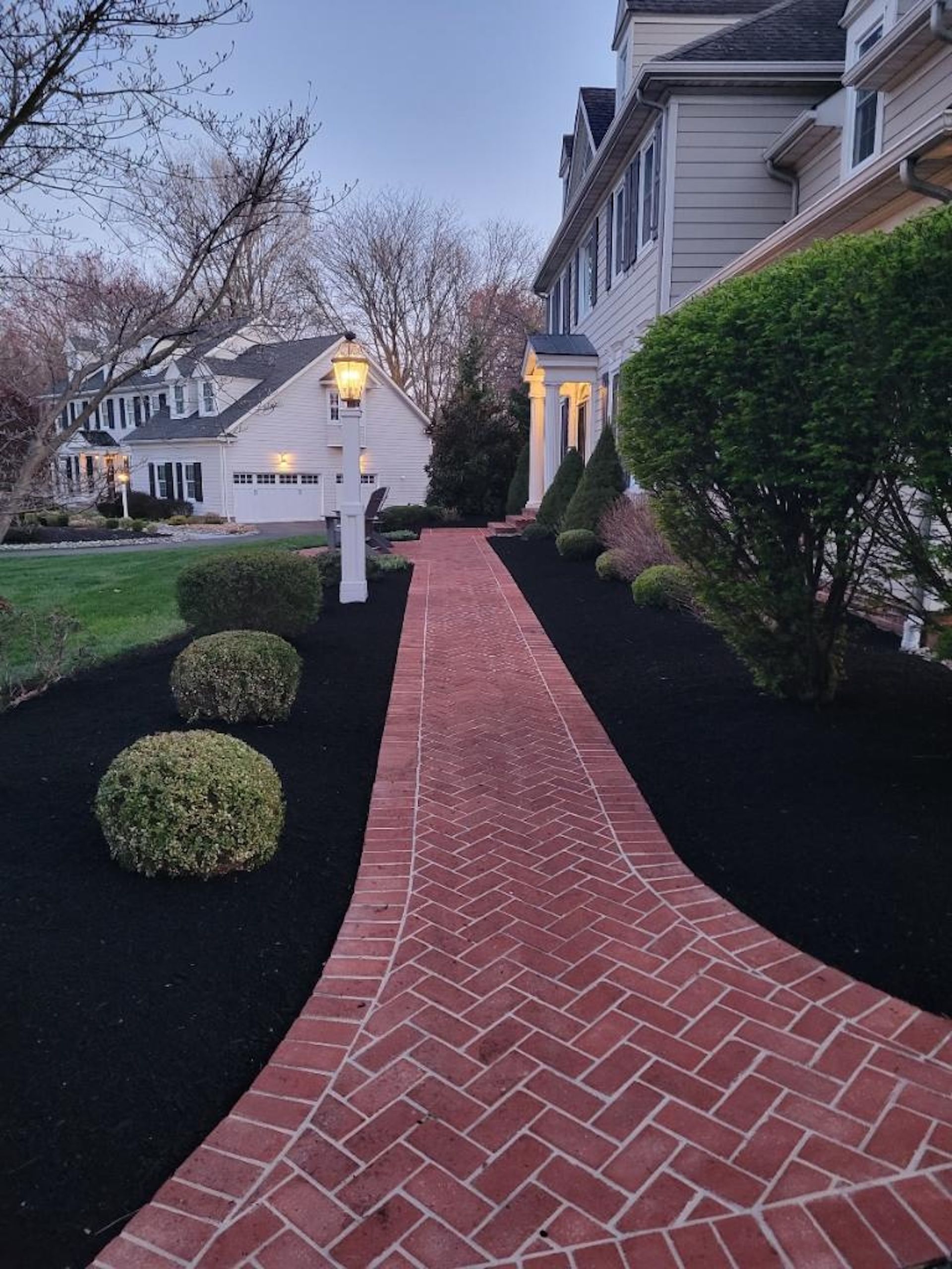 Brick path leading to a white house, flanked by bushes and black mulch.