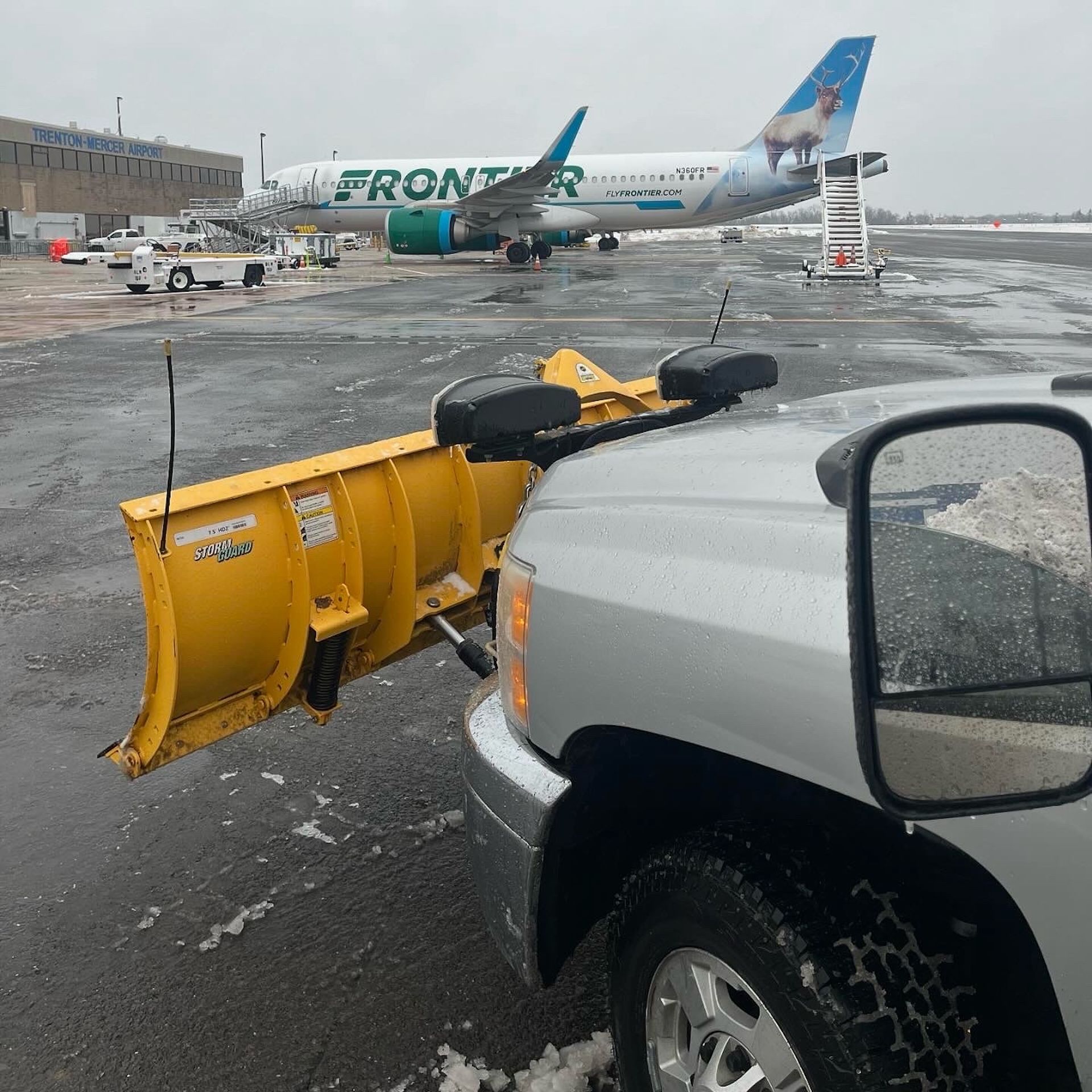 Yellow snow plow in front of Frontier Airlines plane at an airport during snowy conditions.