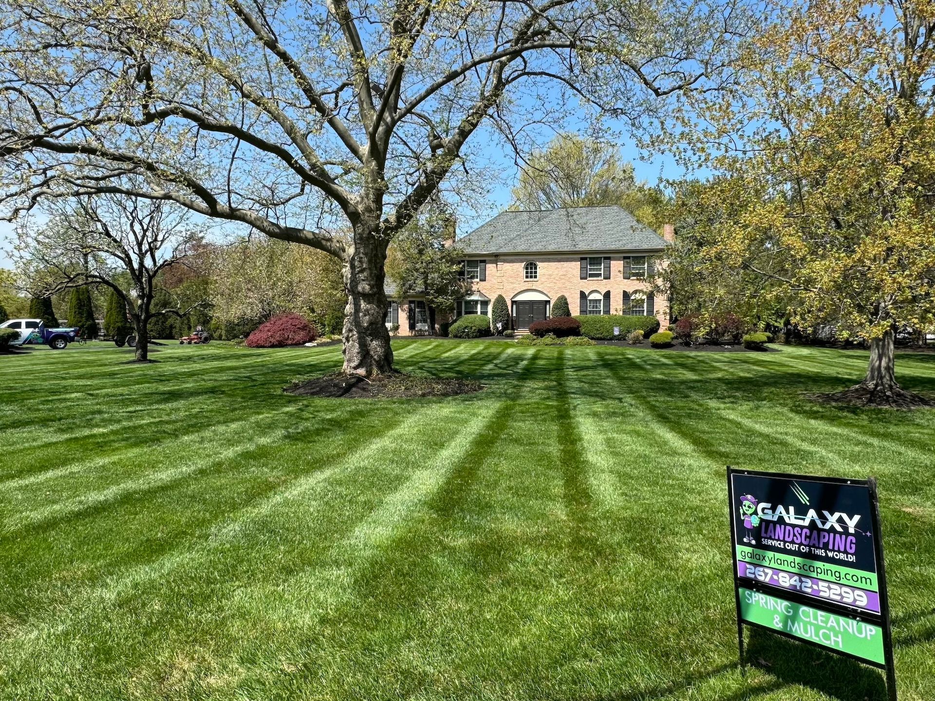 Lawn mowed with stripes in front of a large brick house on a sunny day. A sign for a lawn care company is in view.