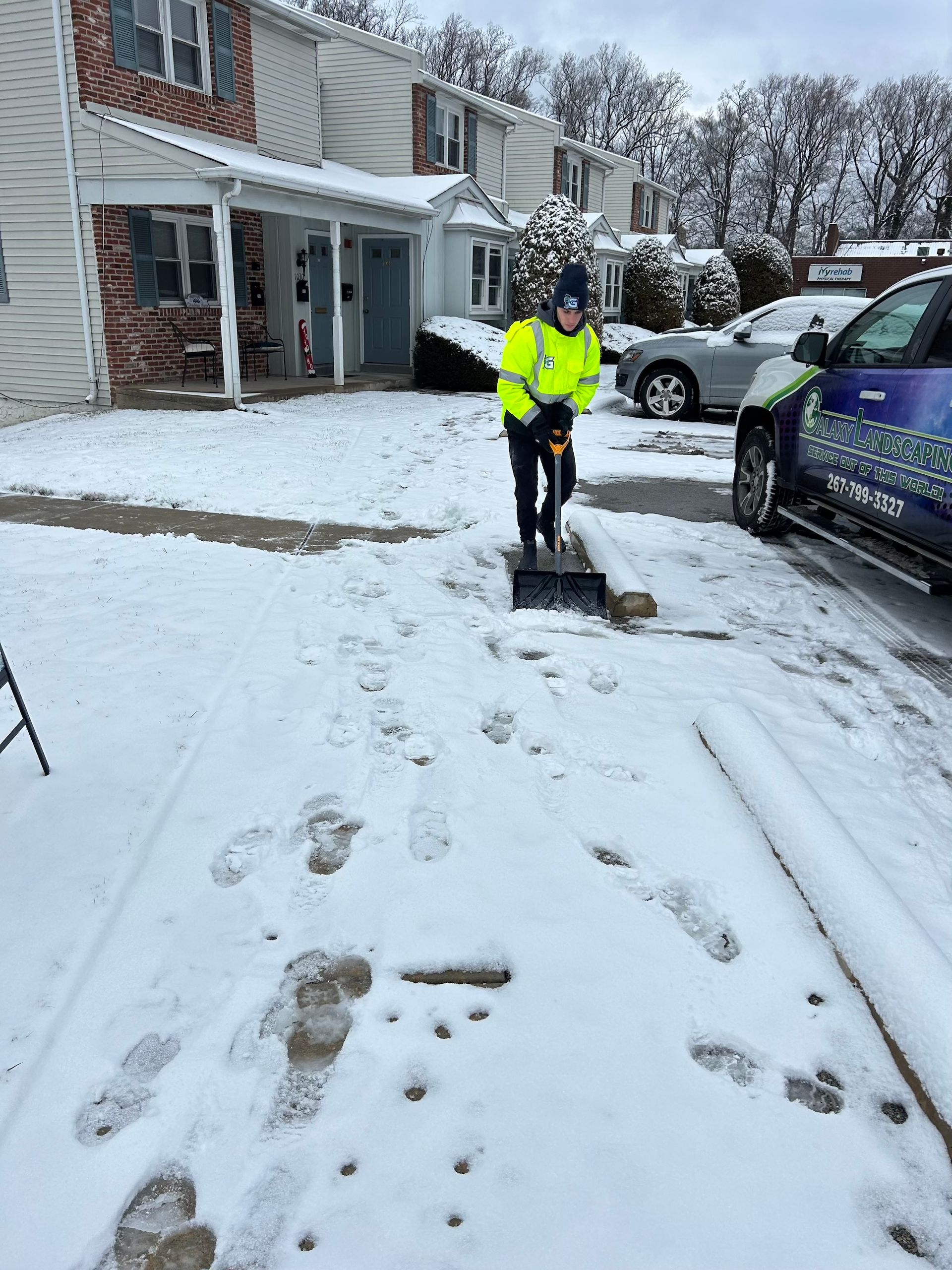 Person in a neon vest shovels snow from a driveway in front of townhouses. Cars are parked nearby.