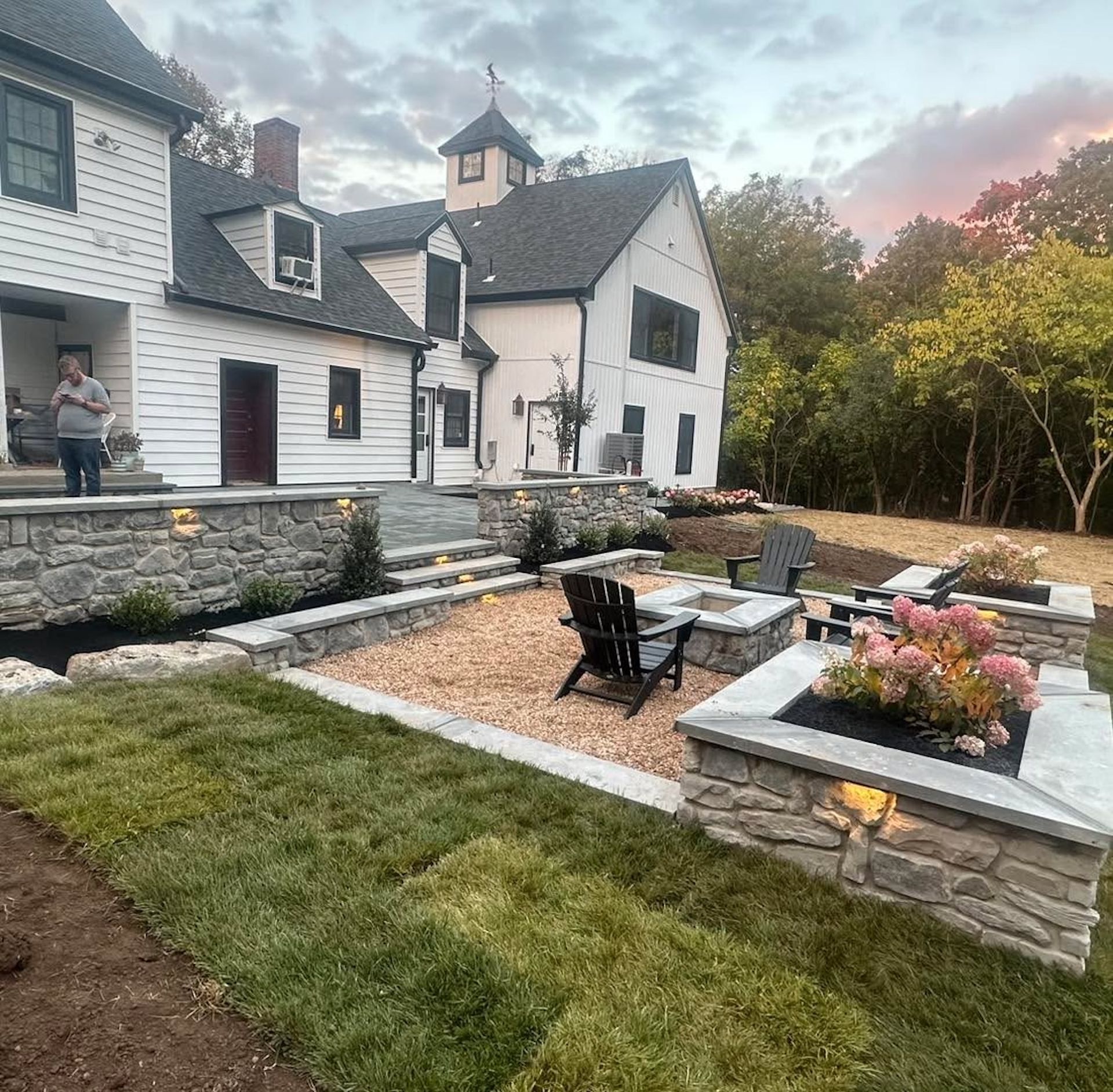 White house with stone patio, fire pit, and landscaping. A person stands on the porch.