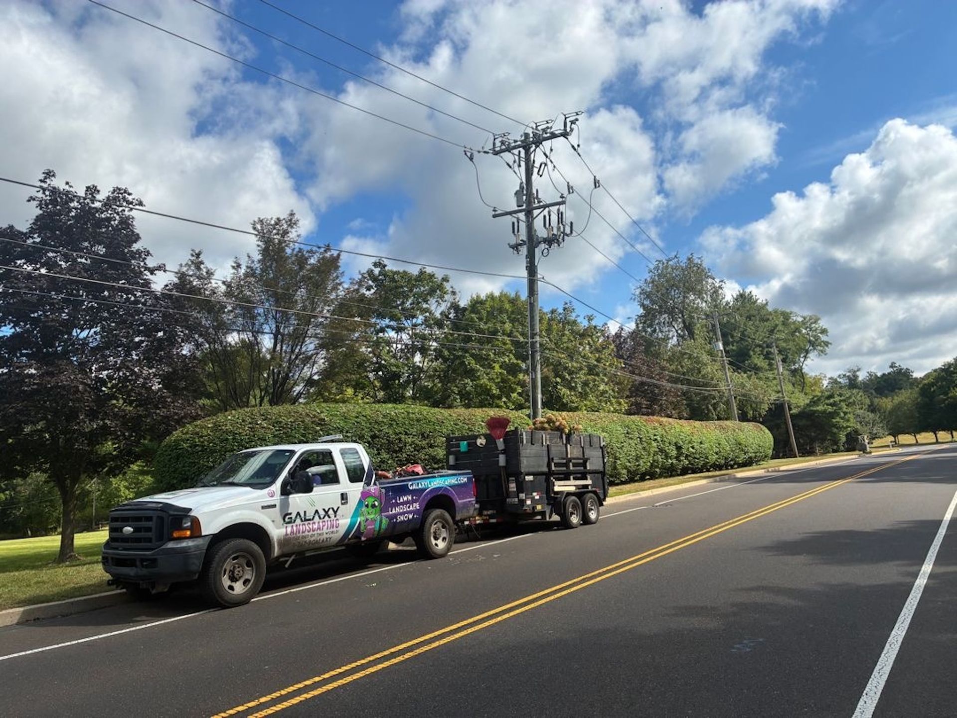 White work truck and trailer parked on roadside, near power lines and trimmed bushes. Bright sky with clouds.