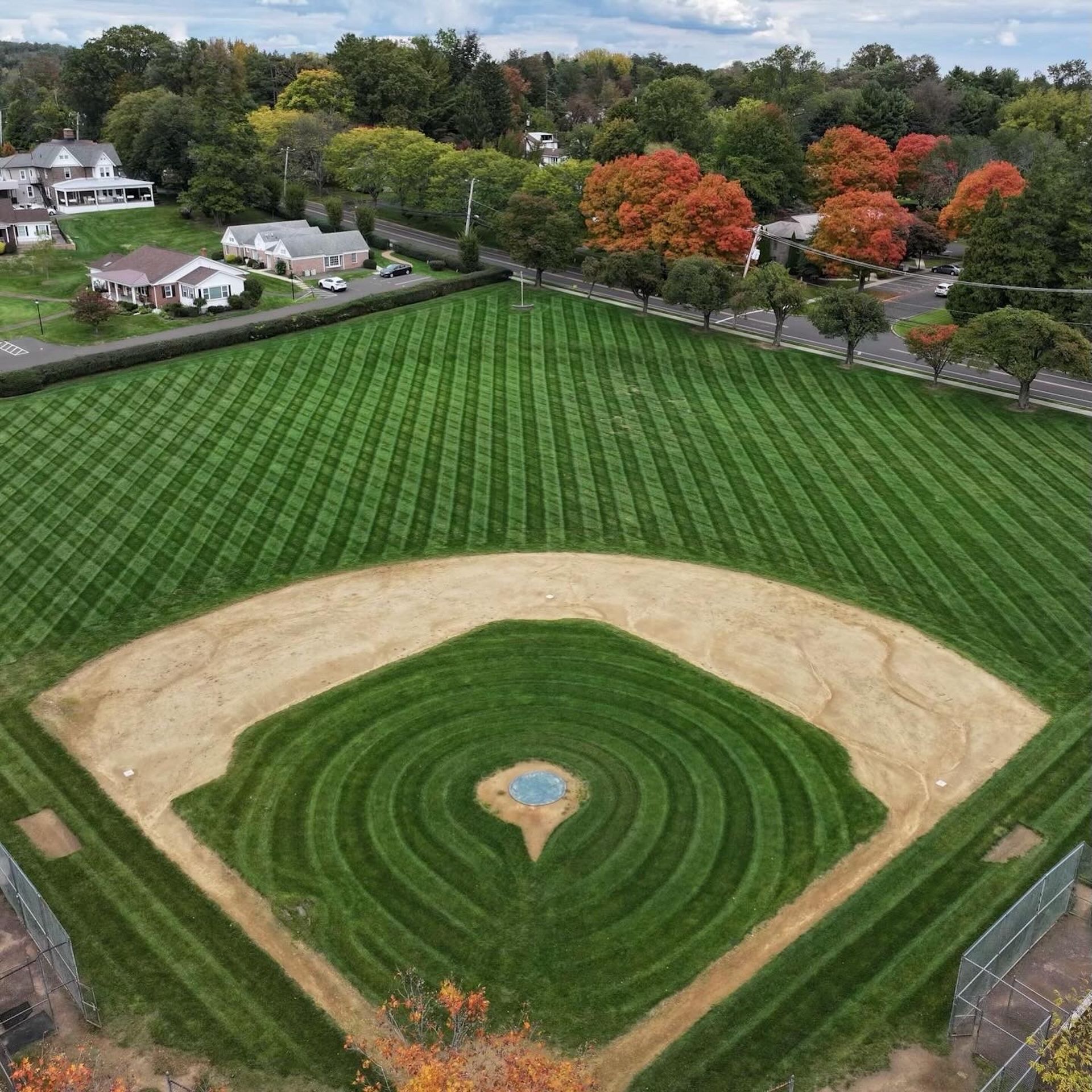Baseball field with meticulously striped green grass, tan infield, and trees with red leaves in autumn.