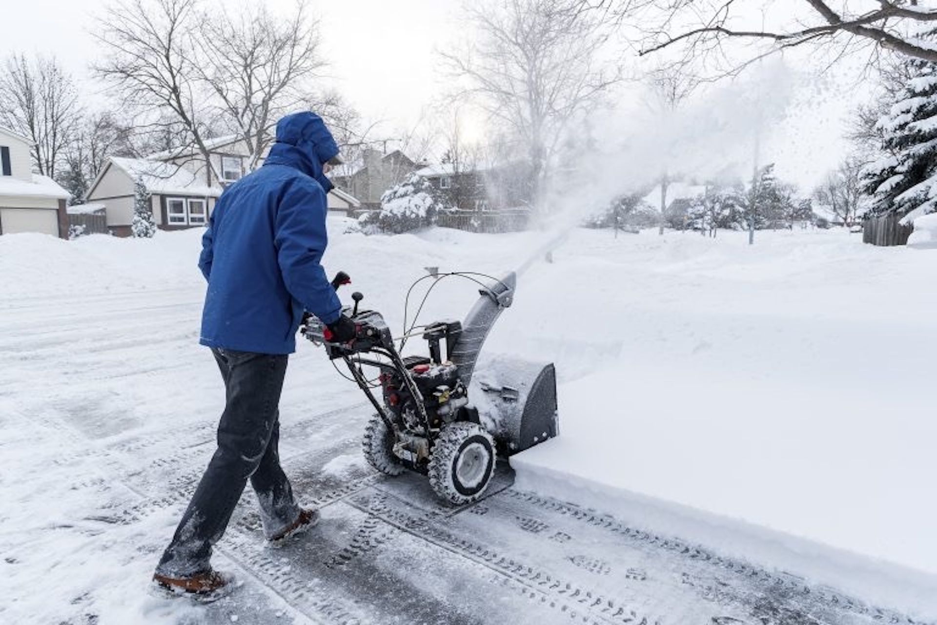 Person in blue coat using snow blower to clear snow from a driveway on a winter day.