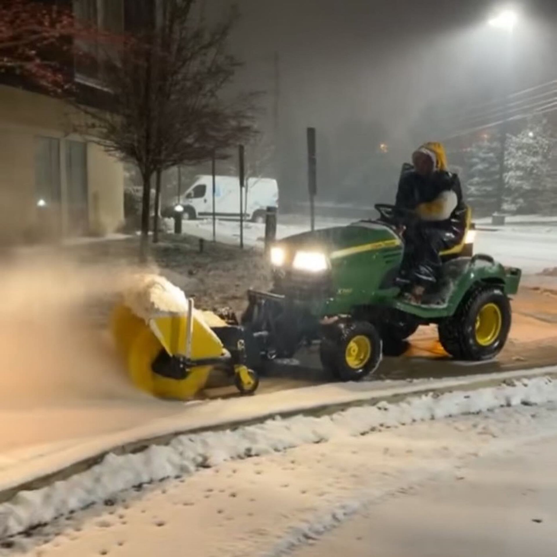 John Deere tractor with snow blower clearing a snow-covered sidewalk at night.