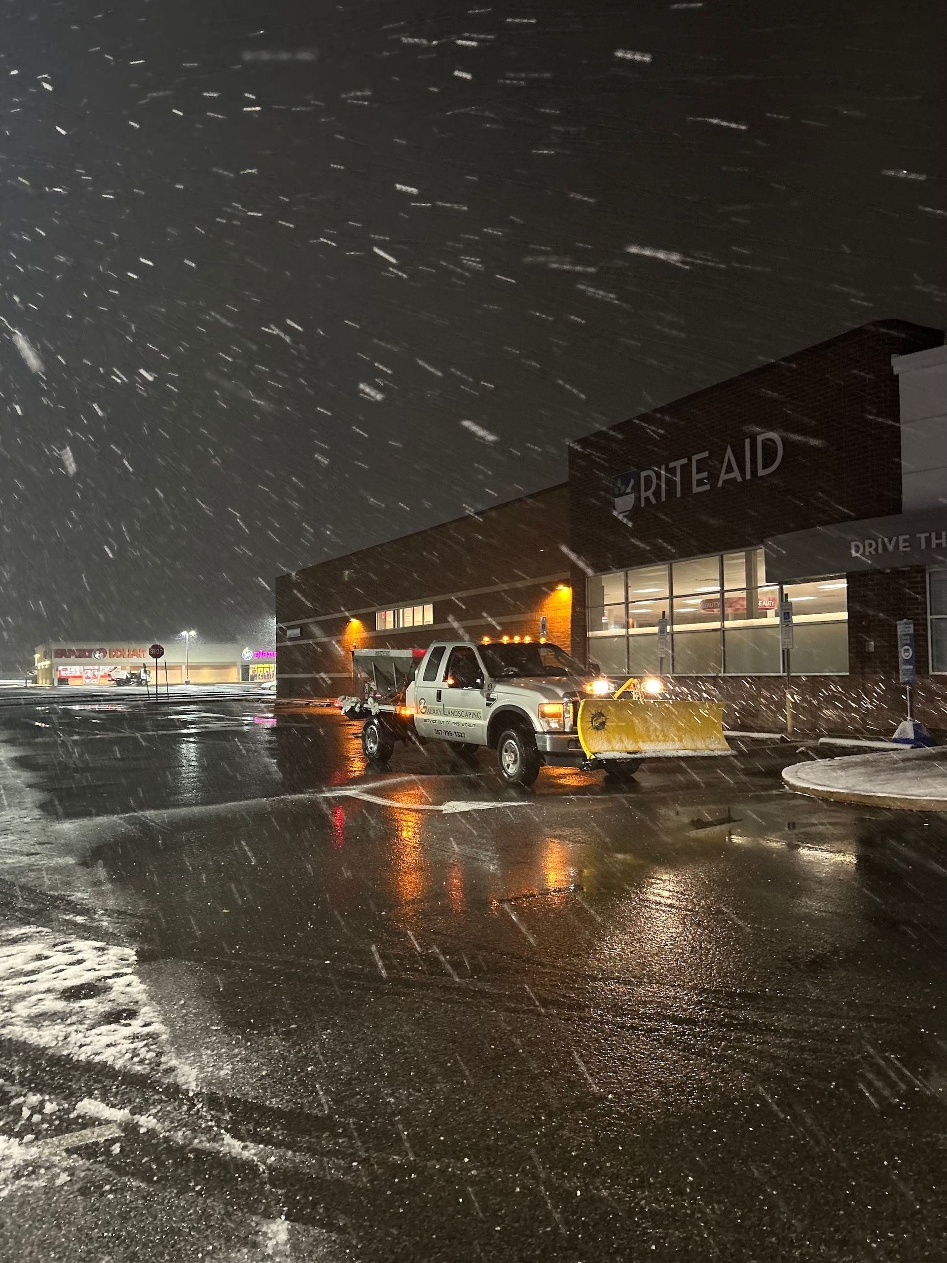 Snowplow in front of a Rite Aid store at night, with snow falling.