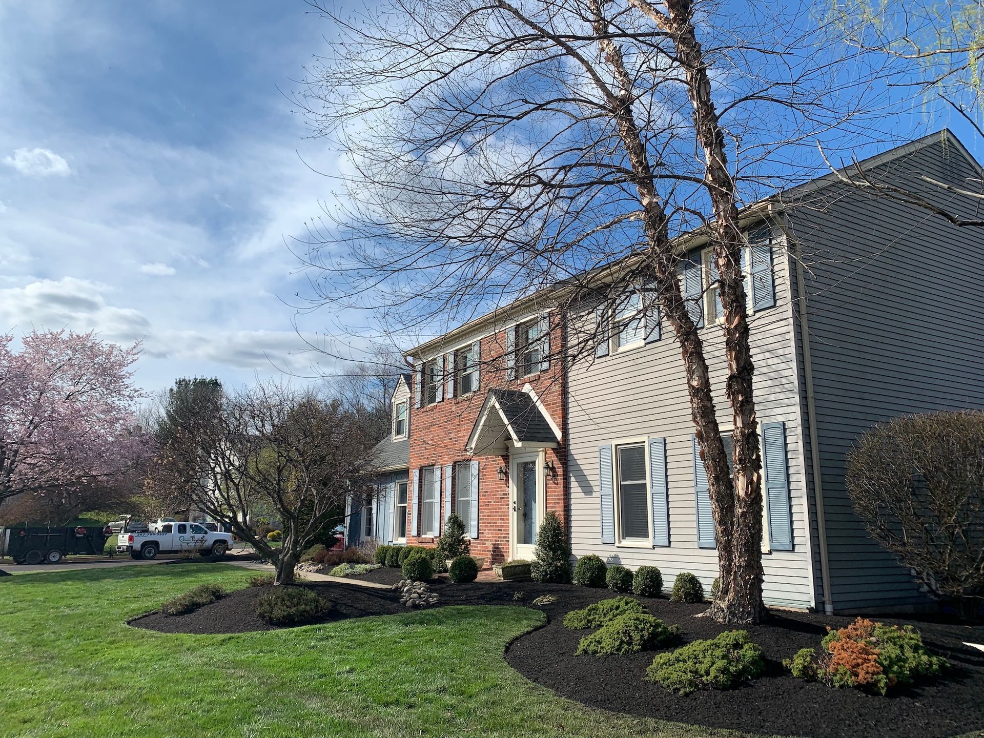 Two-story townhouse with beige siding, blue shutters, and red brick accents. Landscaped front yard with mulch and bushes.