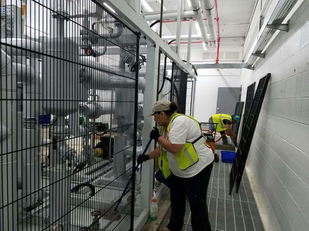 A woman in a yellow vest is cleaning a fence in a factory.