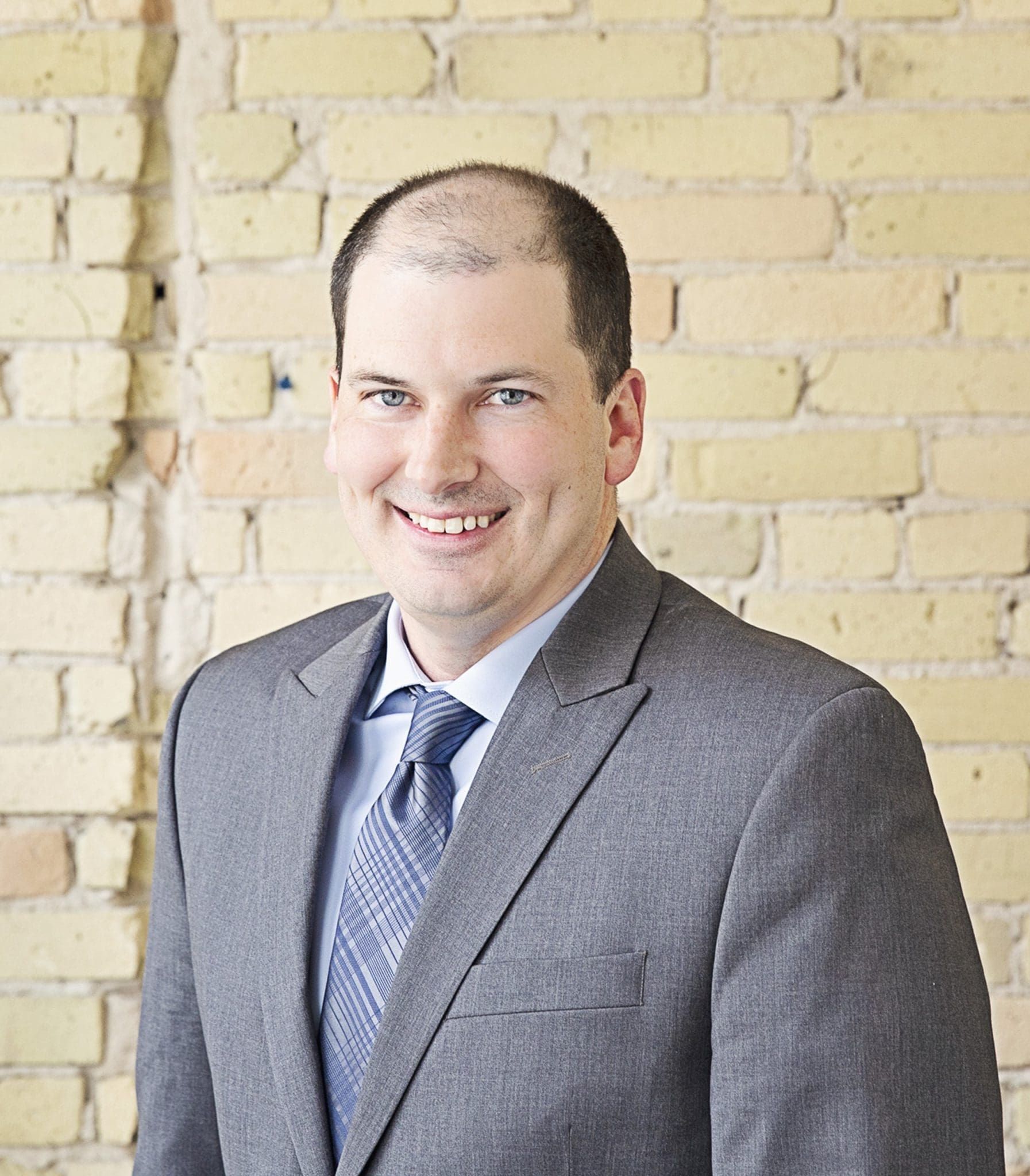 Man in gray suit smiles in front of a brick wall.