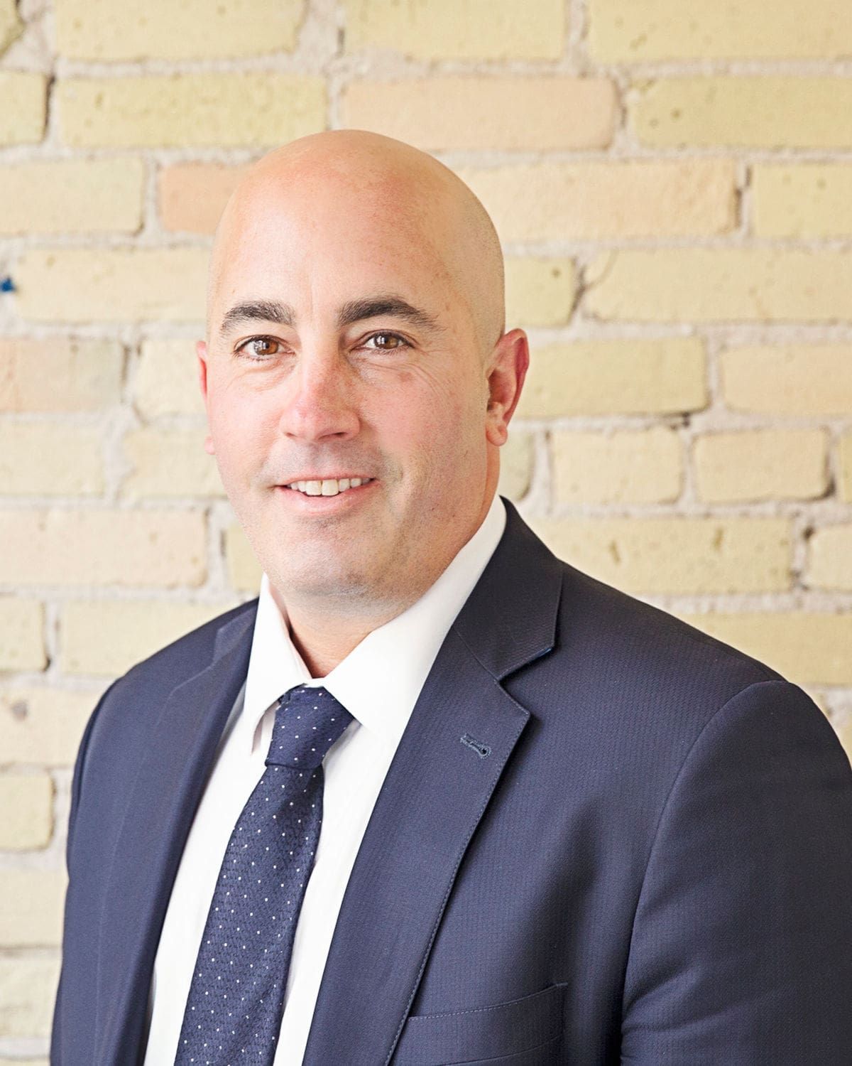 Bald man in a suit smiles in front of a brick wall.