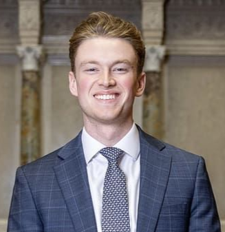 Man in a blue plaid suit and patterned tie smiles; background pillars in a building.