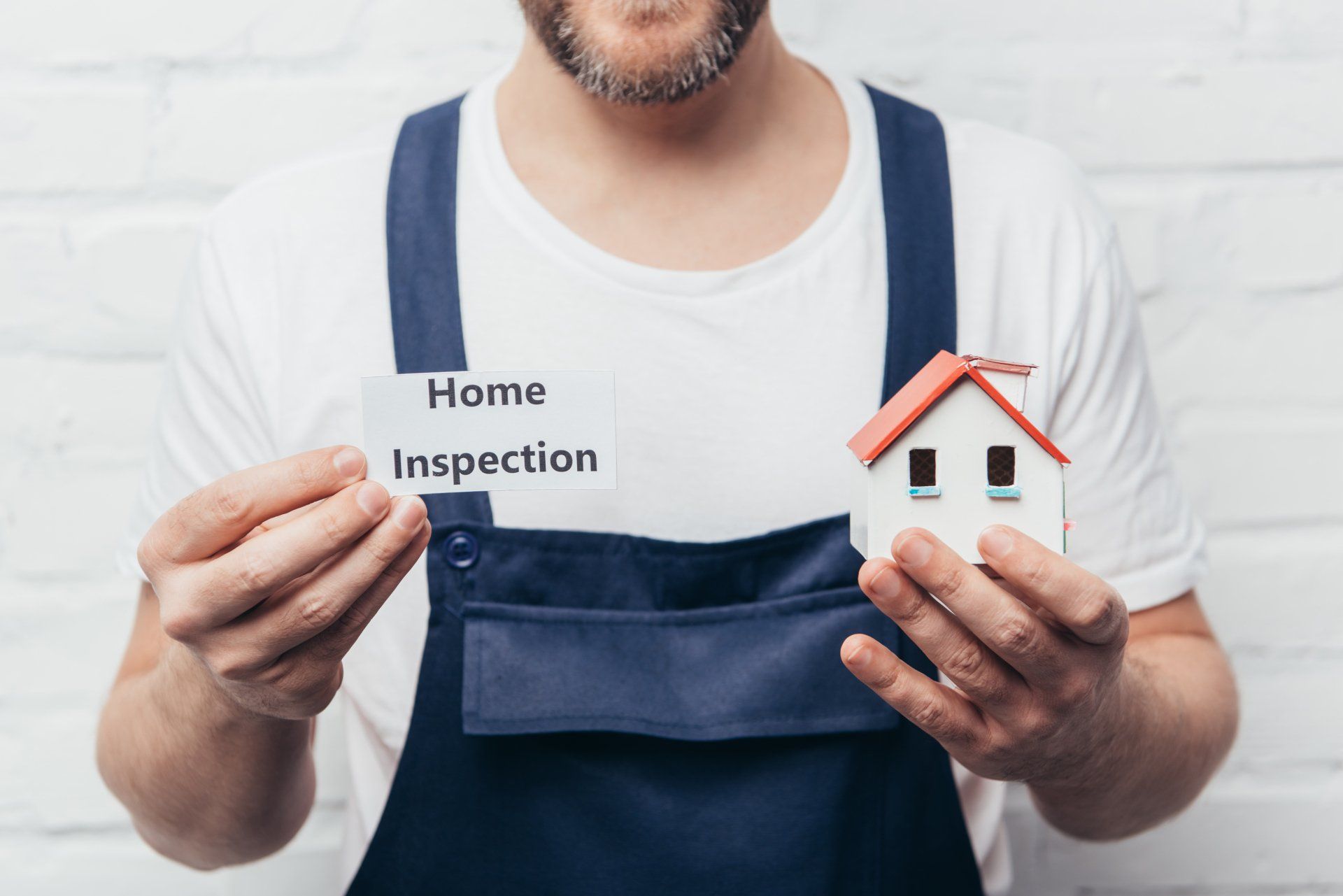 Man i coveralls holding printed square with home inspection in one hand and a house in the other hand