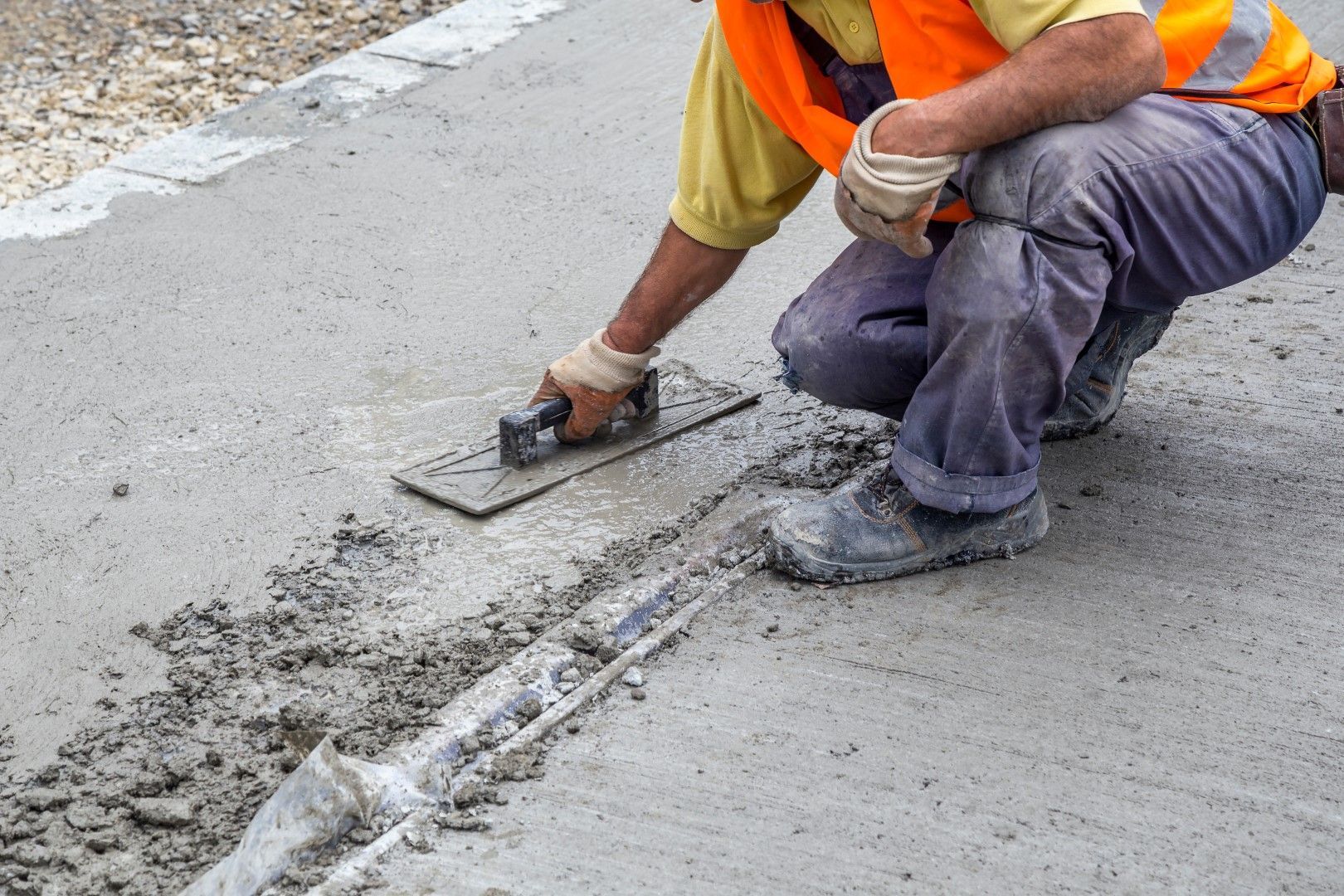 A man is kneeling down and using a trowel to spread concrete on the ground.