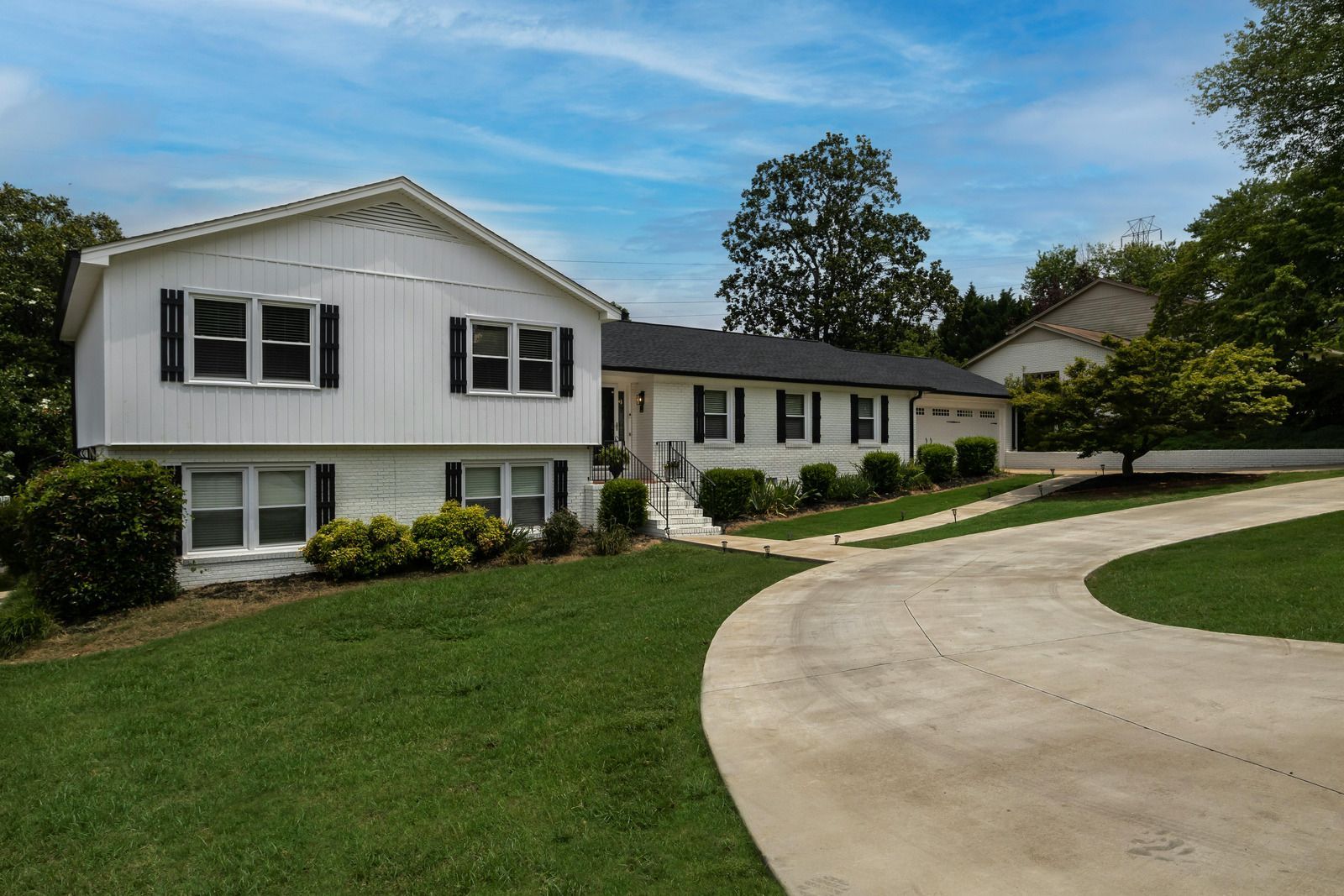 White two-story house with black shutters, driveway, and lush green lawn under a partly cloudy sky.