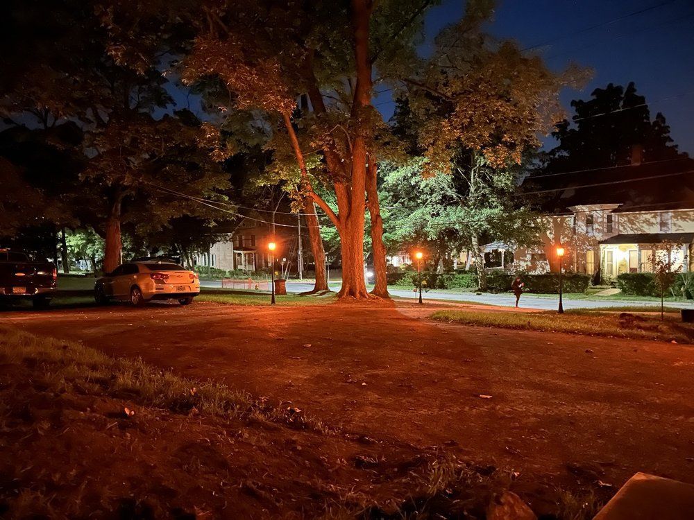 Night scene: Street with trees, lampposts, and a stone house, illuminated under a dark blue sky.