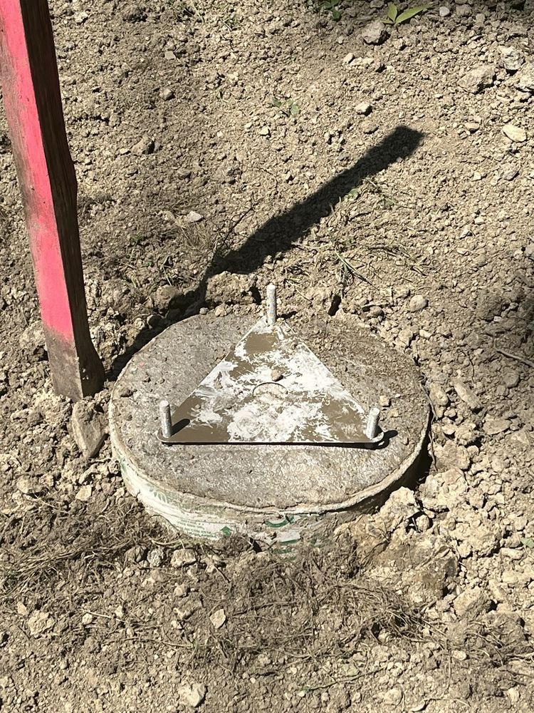 Concrete lid with metal triangle, in dirt, next to a red pole, casting a shadow.