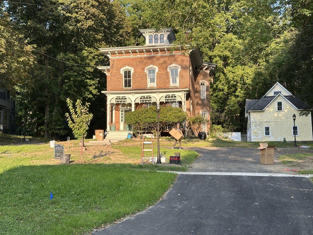 Red brick house with porch and small yellow building. Driveway and grass in foreground.