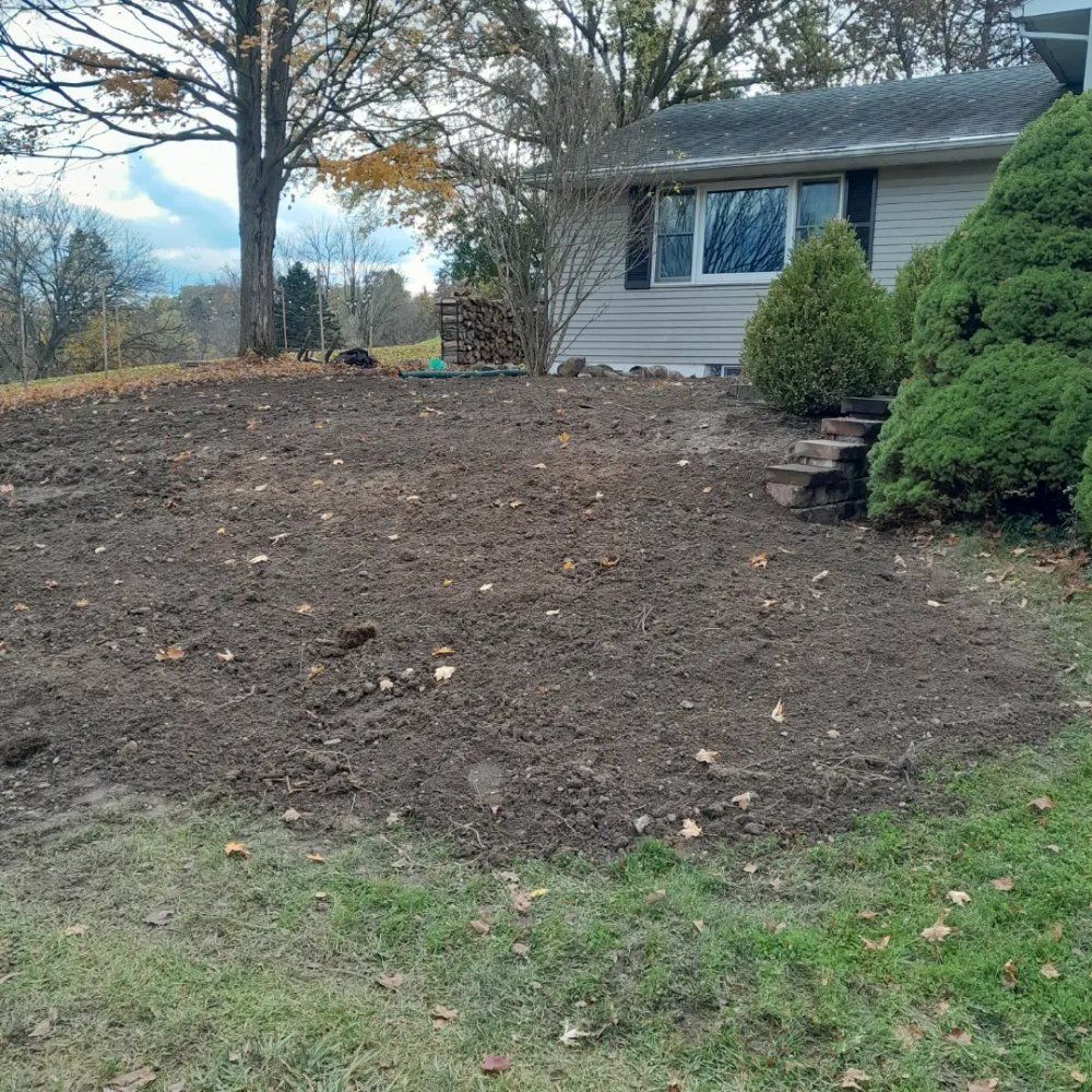 A cleared dirt patch in front of a house, with grass, bushes, and a pile of wood in the background.