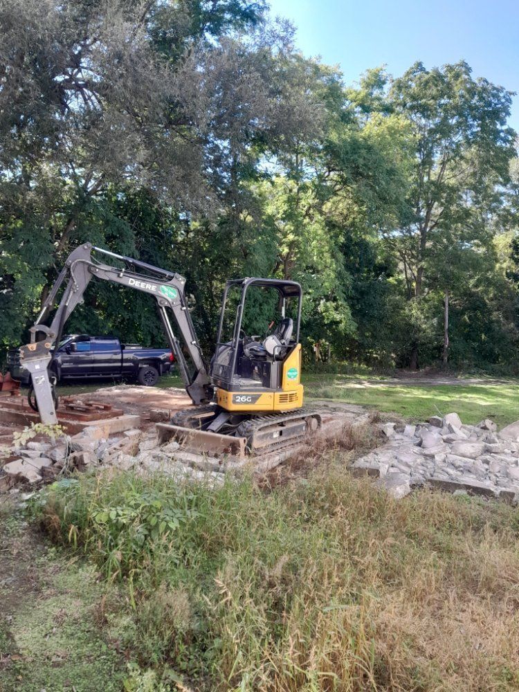 A yellow and black excavator working on a construction site; overgrown grass, trees, and blue sky in the background.
