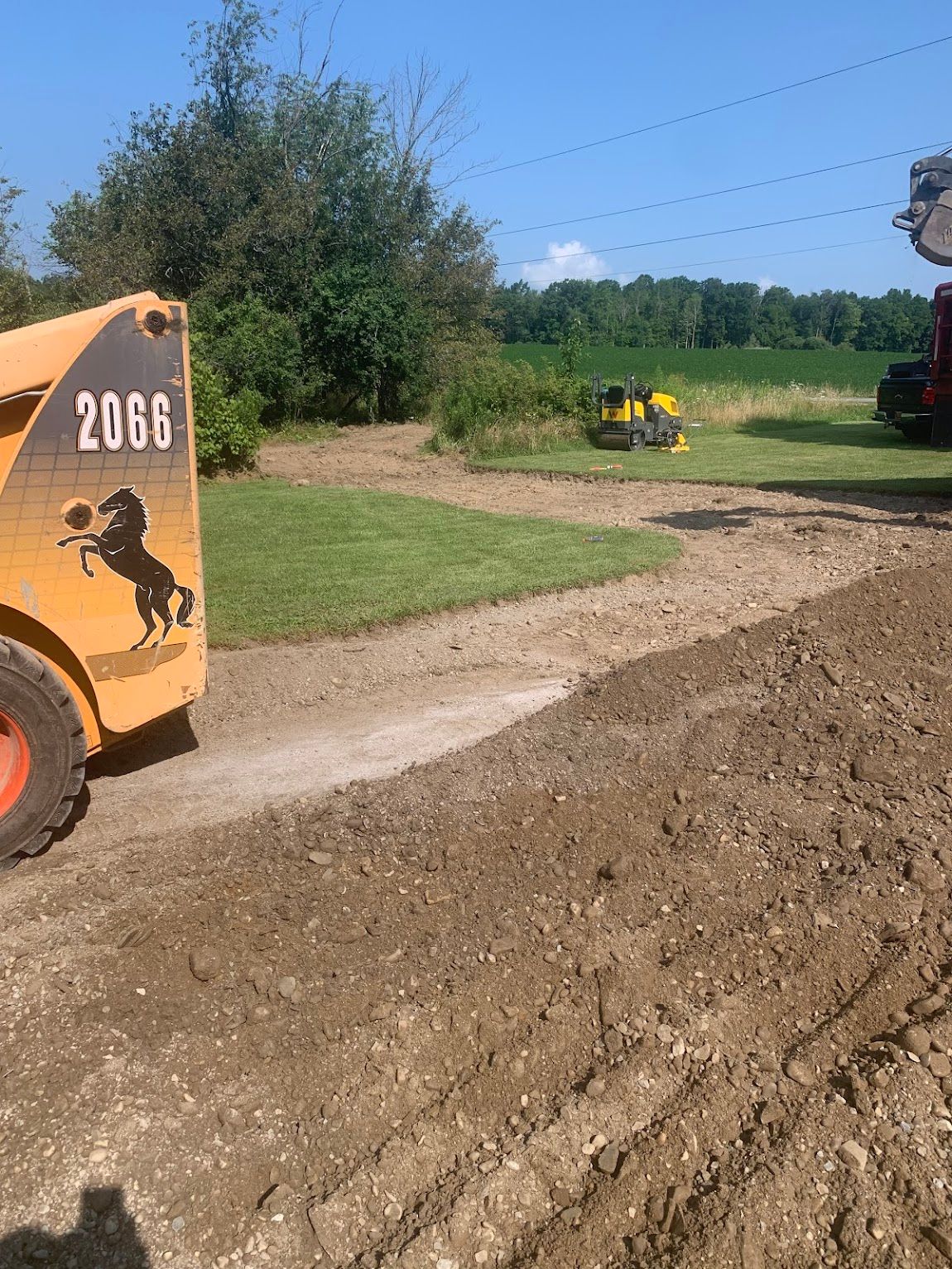 A skid steer, roller, and truck are on a dirt path in a grassy area near trees.