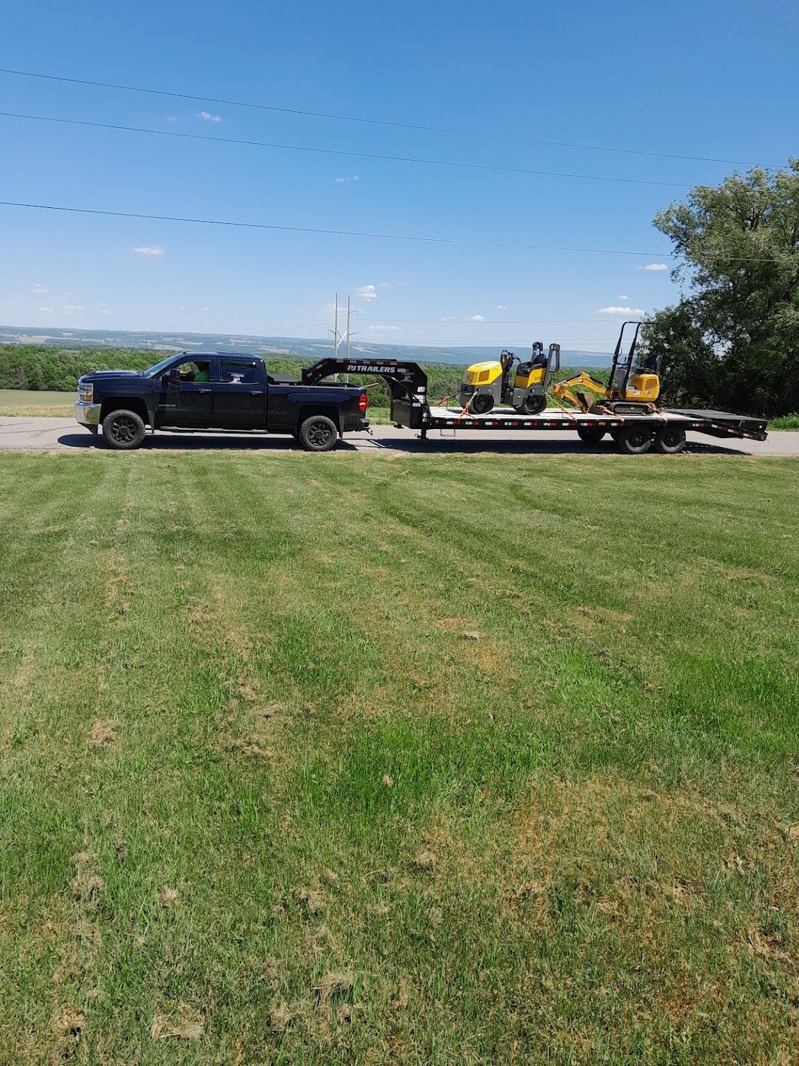 Black truck towing trailer with yellow construction equipment on a green lawn, under blue sky.