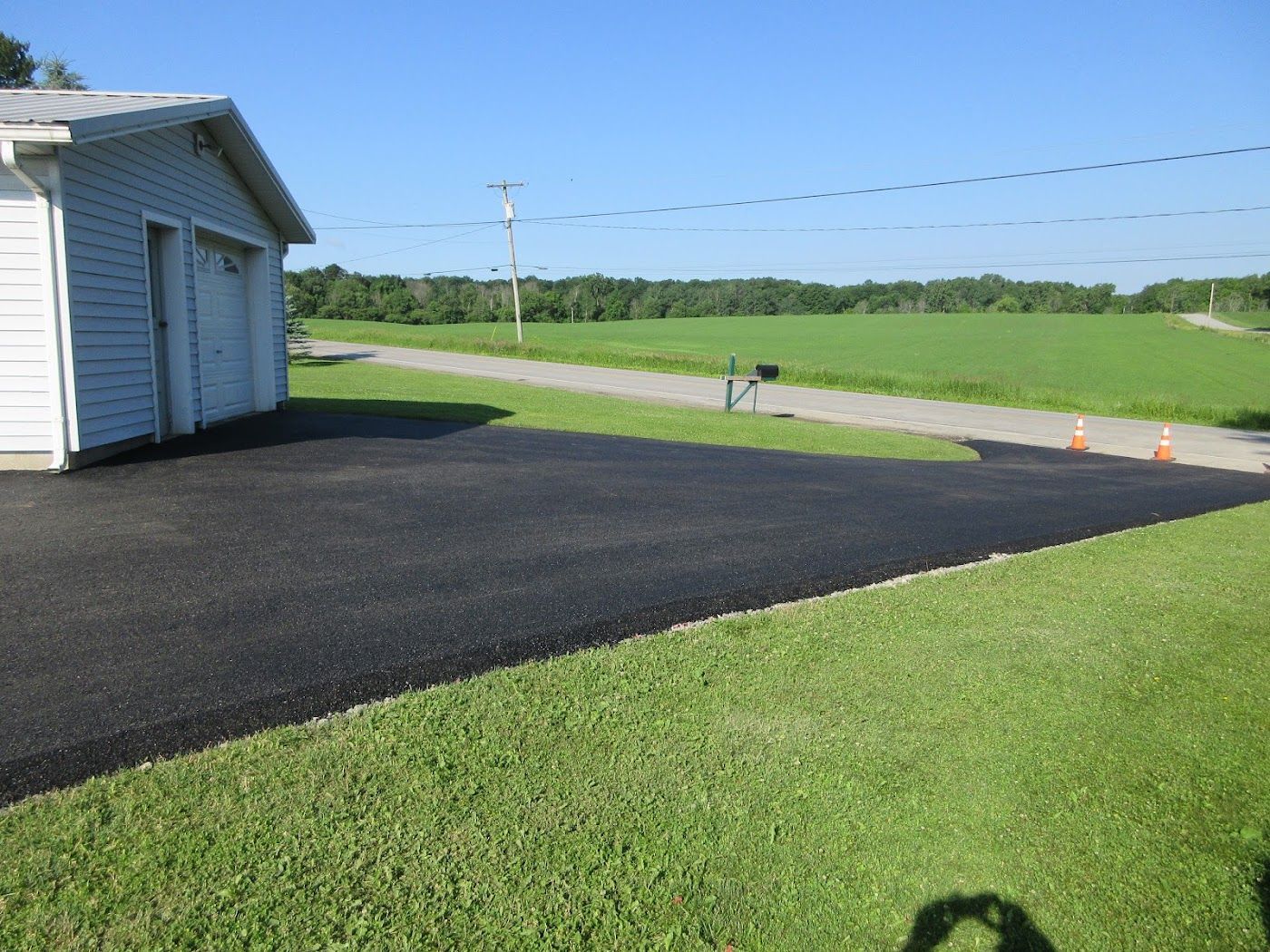 Asphalt driveway next to a white garage, green grass, and a rural landscape under a blue sky.