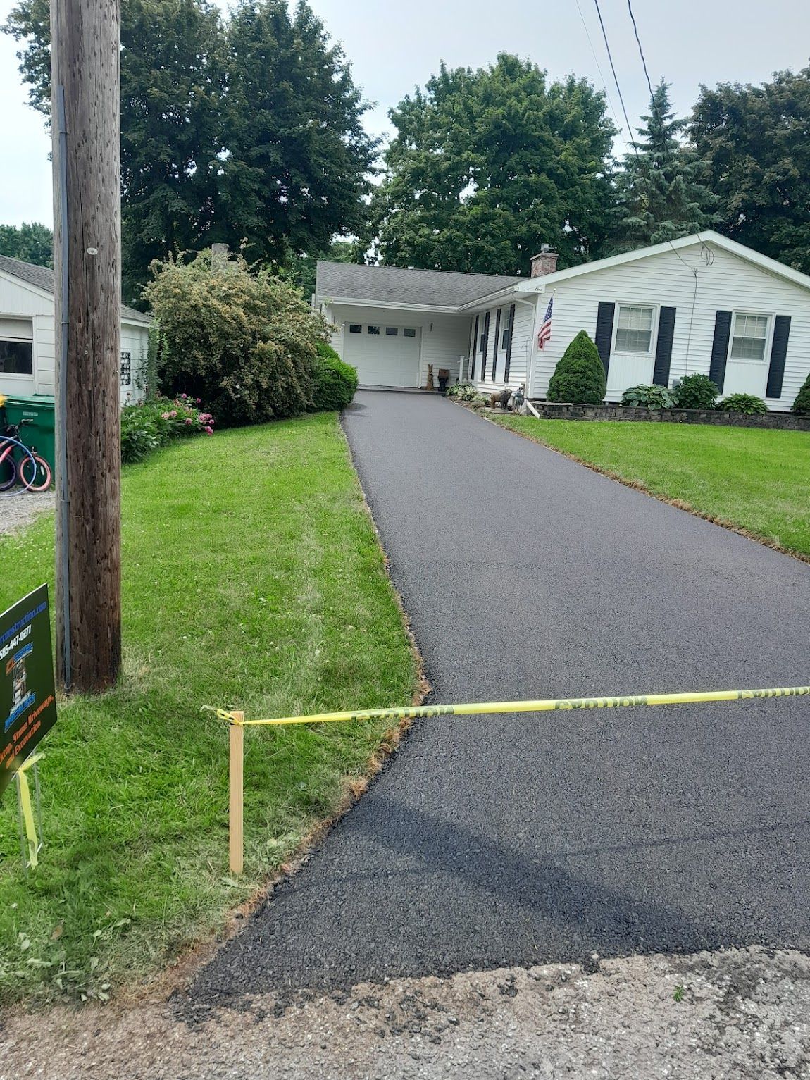 Newly paved asphalt driveway leading to a white house with a garage, green lawn, and utility pole.