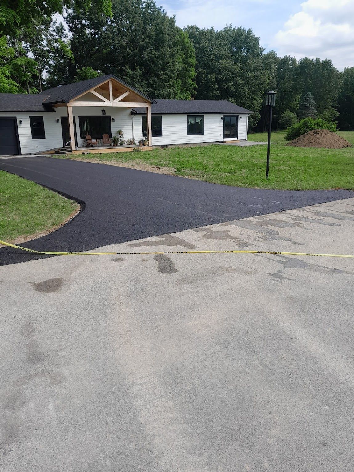 New black asphalt driveway curves to a white house with a porch and trees.