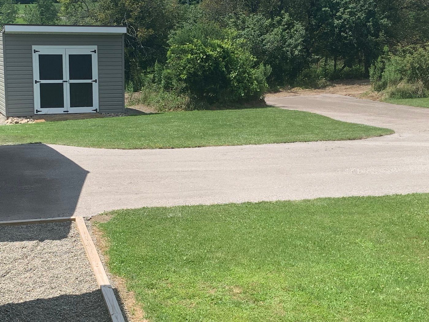 Gray shed with white-framed windows, beside a concrete path and lawn, with trees in background.