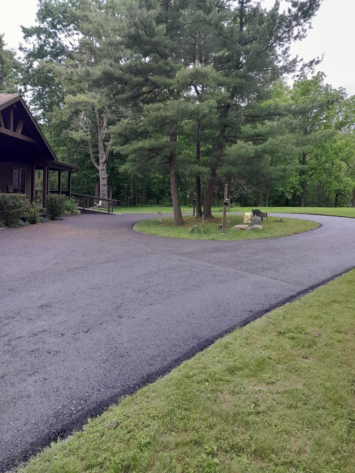 Paved driveway curves around a tree on a grassy lawn, leading to a cabin in a wooded area.