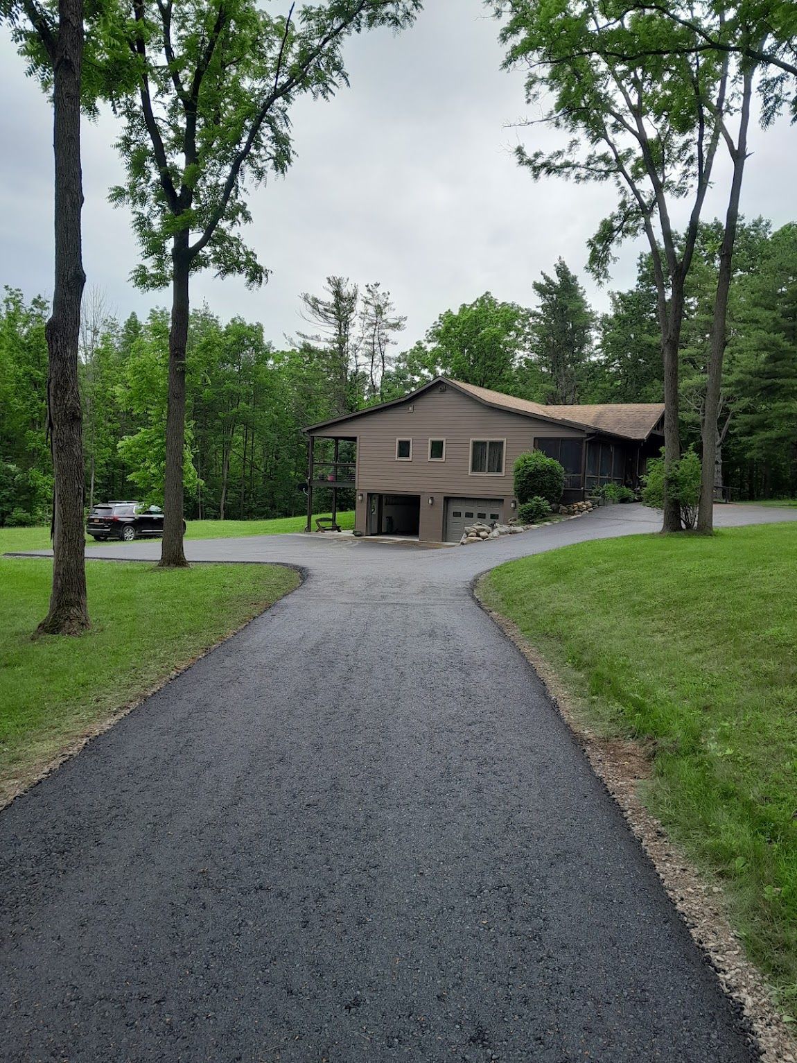 Asphalt driveway leads to a two-story house with a garage, surrounded by trees and green grass on a cloudy day.