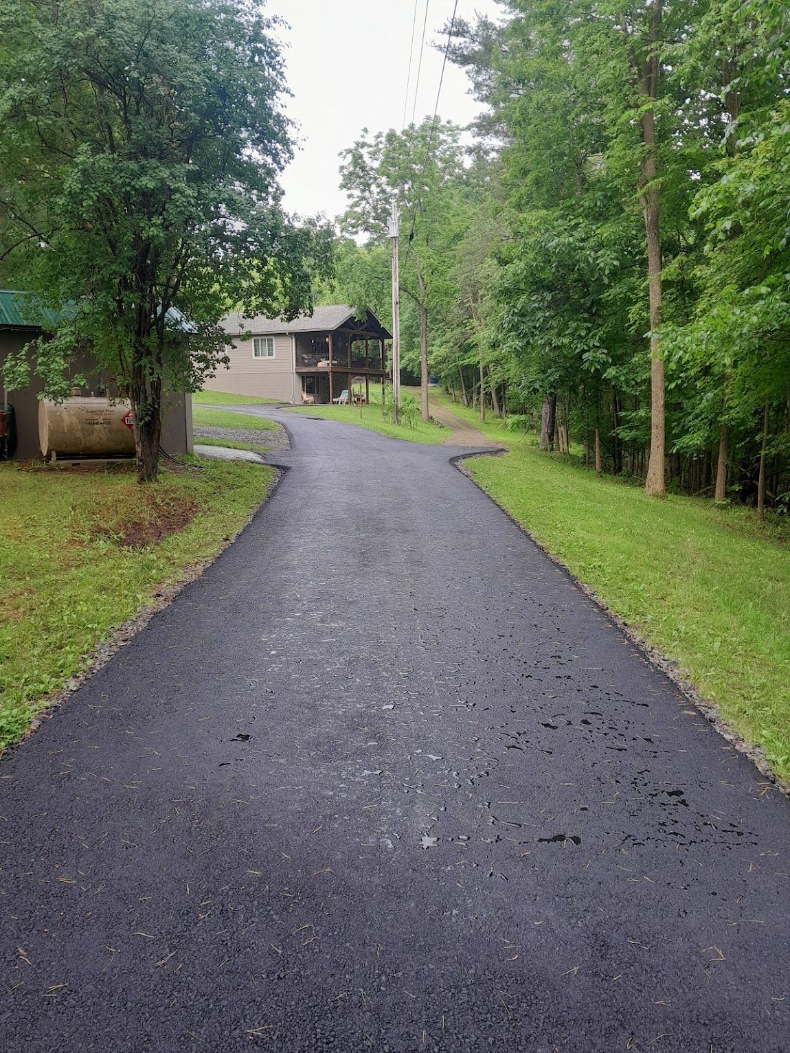Asphalt driveway leading to a cabin in a wooded area with green grass and trees.