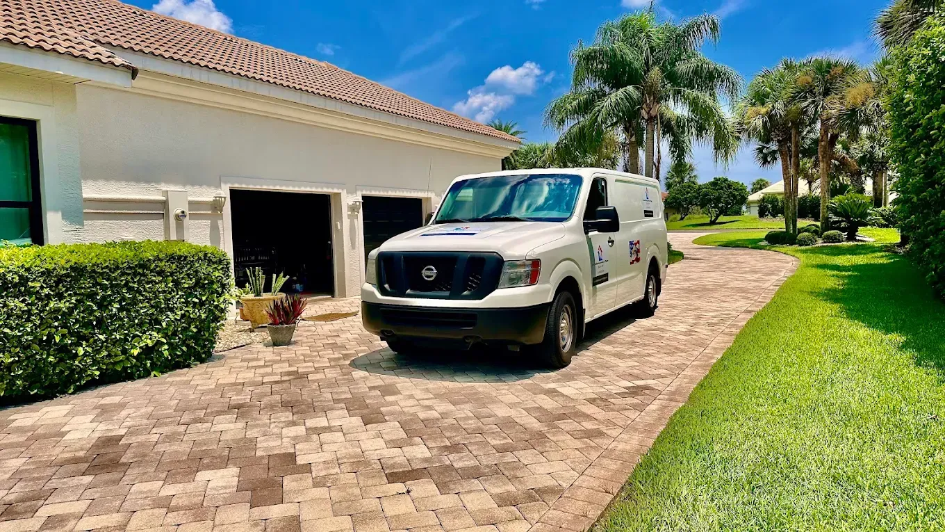 A white service van parked on a brick driveway in front of a house with a green hedge and palm trees under a blue sky.