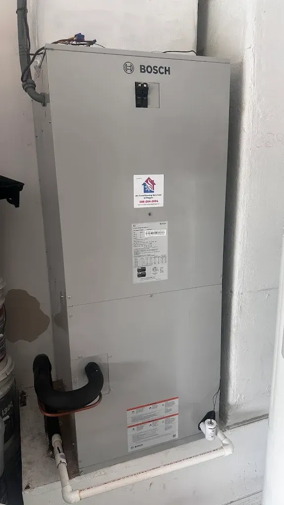 A gray Bosch air handler unit stands indoors against a wall, featuring a circuit breaker, HVAC labels, and drainage pipes.