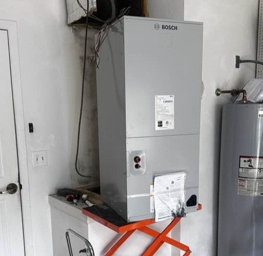 A gray Bosch air handler unit rests on an orange scissor lift in a residential room next to a water heater.