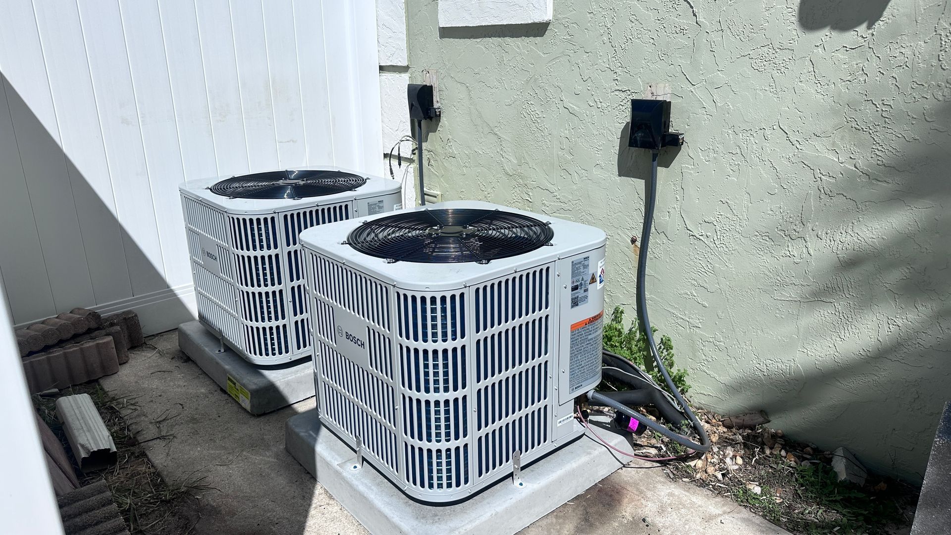 Two white, square residential air conditioning units sitting on concrete pads against a light green exterior house wall.