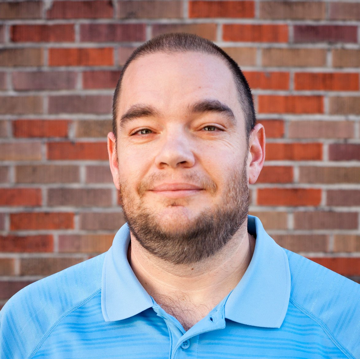 A man in a blue shirt is smiling in front of a brick wall.