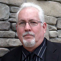 A man with glasses and a beard is standing in front of a stone wall.