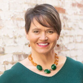 A woman wearing a green sweater and a green necklace is smiling in front of a brick wall.