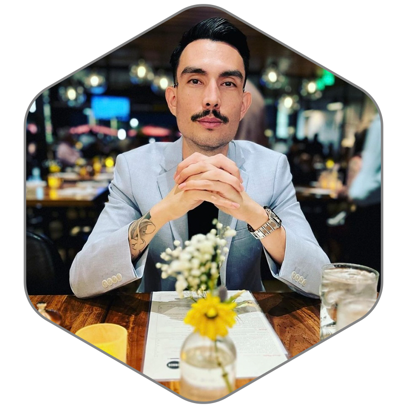 A man with a mustache is sitting at a table with his hands folded.