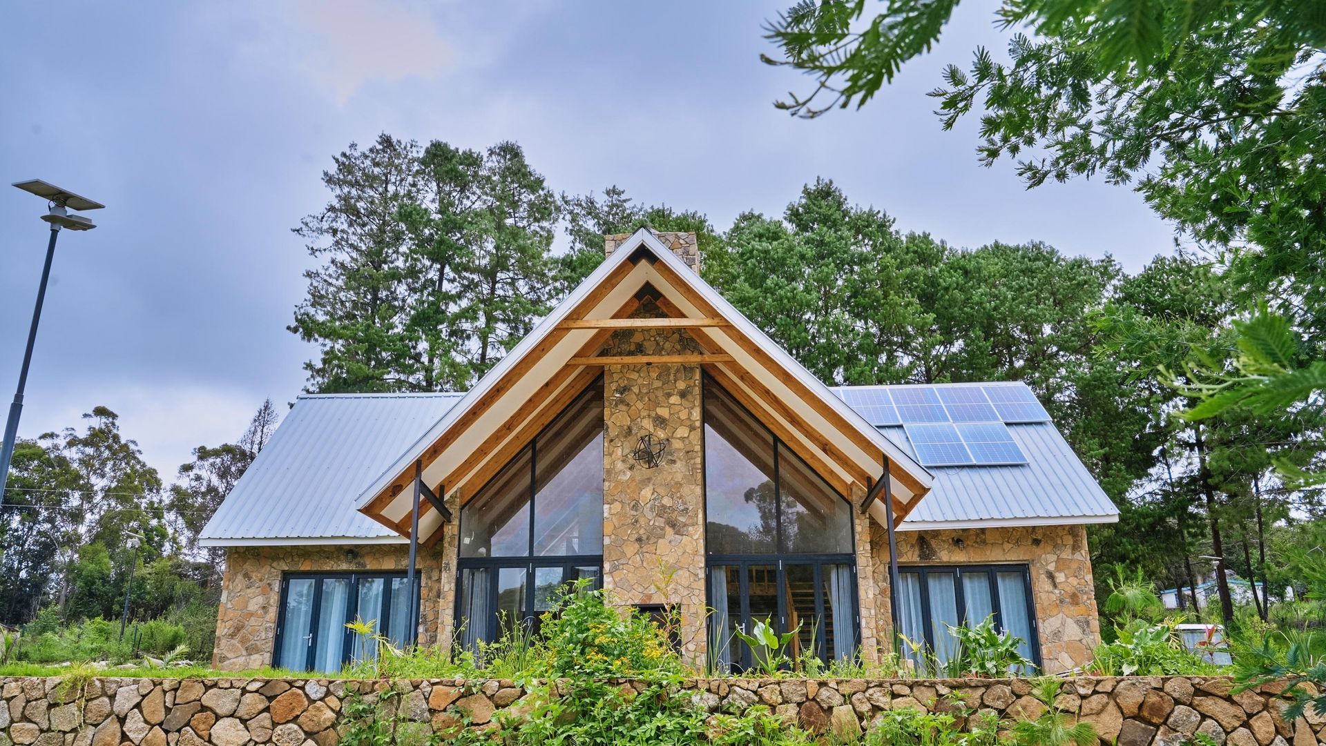 A large stone house with a metal roof and solar panels on the roof surrounded by trees.