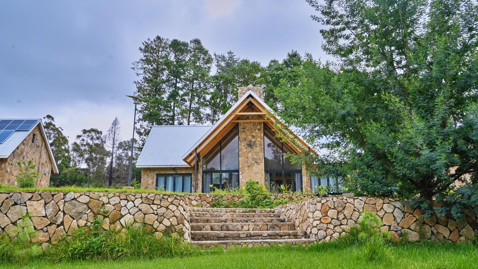 A house with a stone wall in front of it is surrounded by trees.