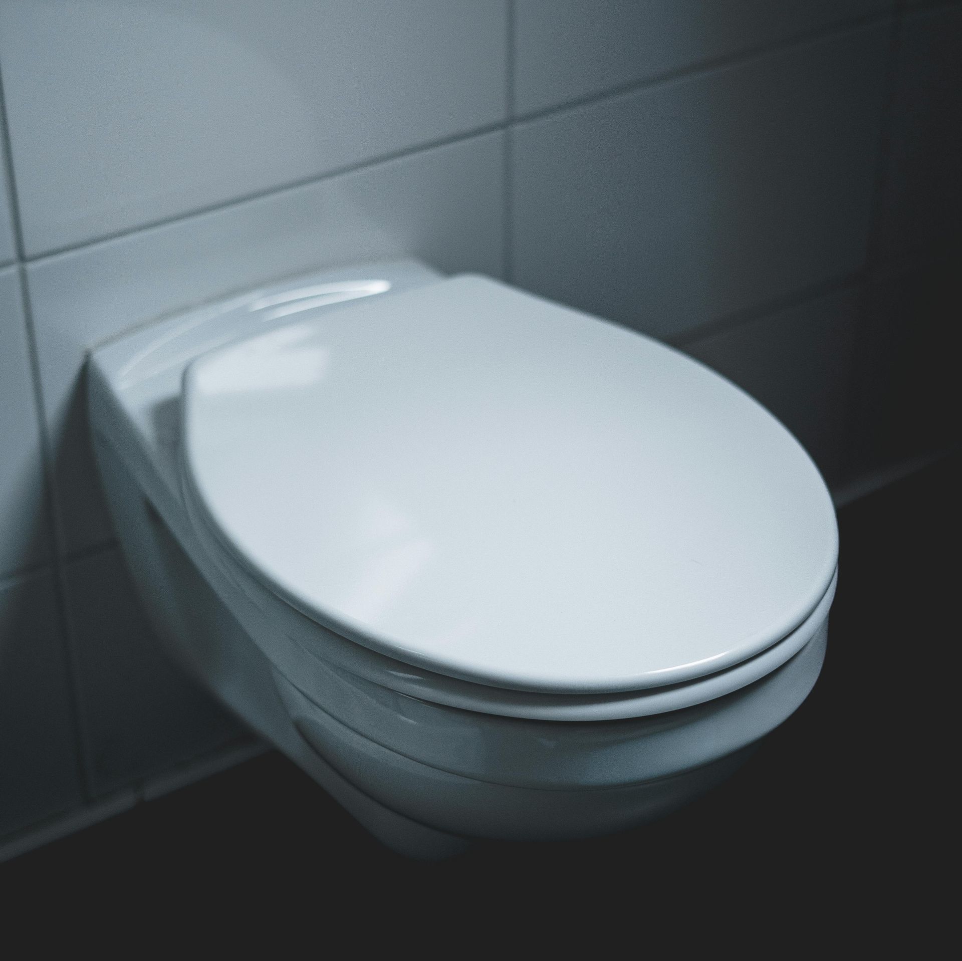 White, wall-mounted toilet with closed lid, set against white tiled wall and dark floor.