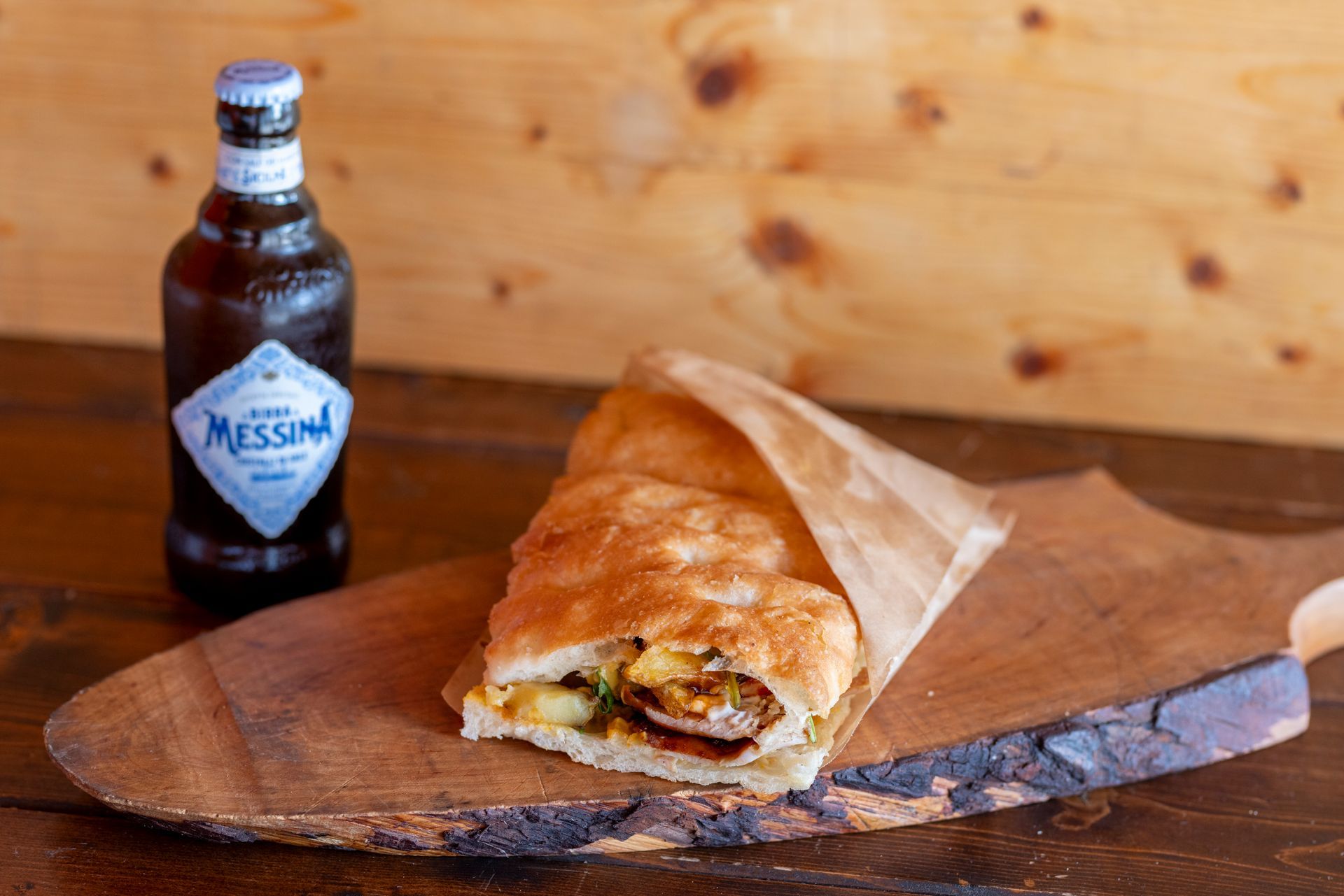 Un panino avvolto nella carta è appoggiato su un tagliere di legno accanto a una bottiglia di birra messina.