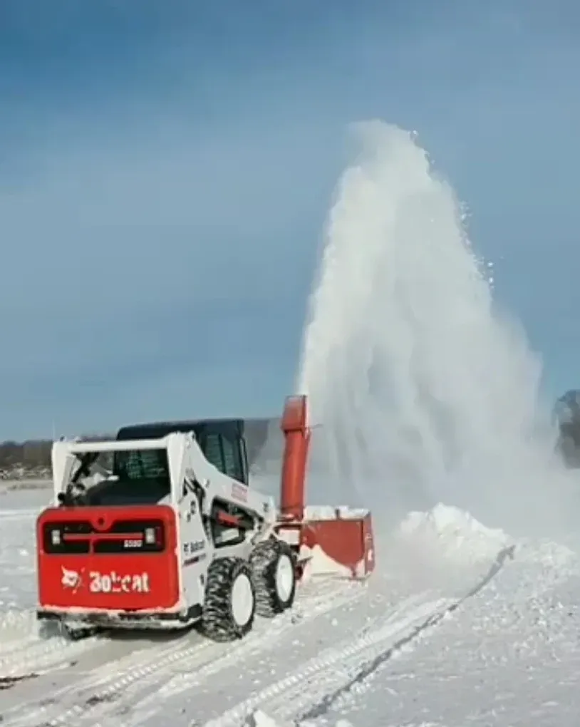 A Bobcat skid-steer loader with snowblower clears snow, ejecting a plume into the air, on a snowy field.