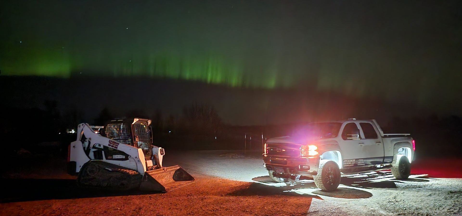 A white pickup truck and a skid steer are illuminated by headlights under green aurora borealis.