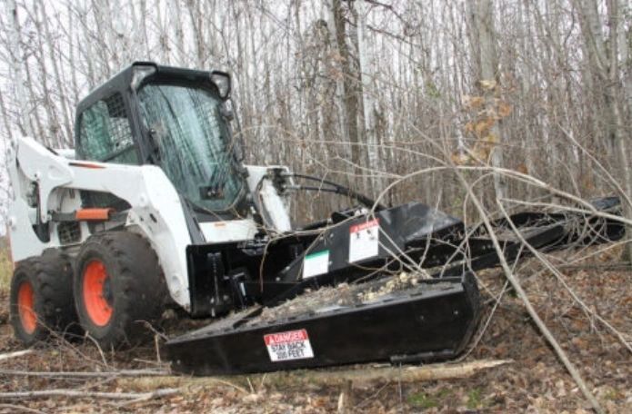 A white Bobcat skid steer with a brush cutter attachment clearing a wooded area.